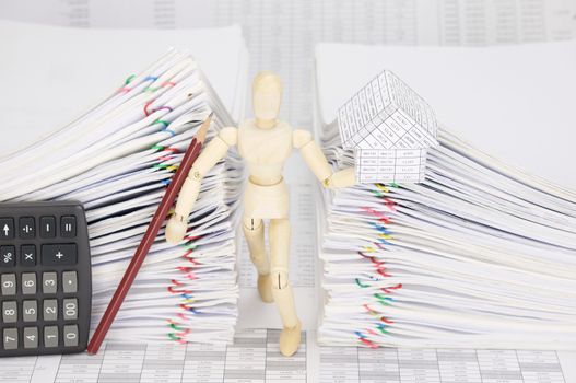 Wooden dummy holding house and pencil with calculator between dual pile overload document of report with colorful paperclip on finance account.