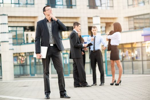 Young businessman standing in front of office building separated from the rest of the business team holding a closed laptop and happily looking into the distance, thinking about business success.