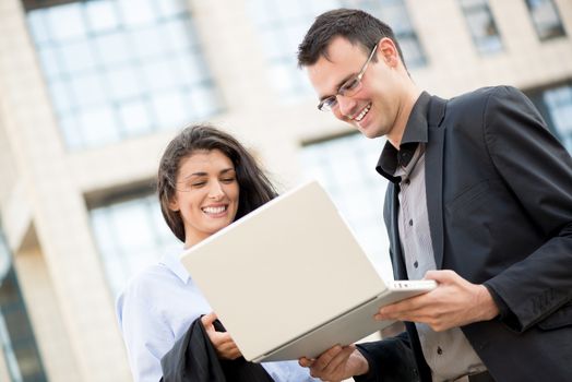 Smiling young businessman and businesswoman in front of office building looking at the laptop that holds a businessman.