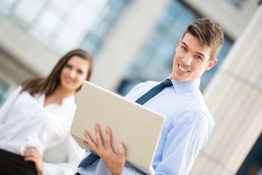 Handsome young businessman with a laptop in front of their office building with his partner in the background looking at the camera.