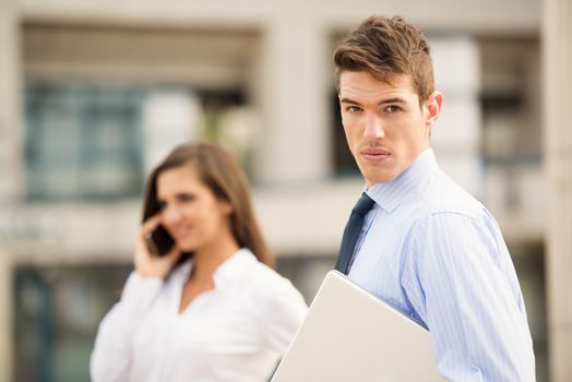 Young business couple standing in front of the building. Businessman carrying a laptop, while his female partner use a mobile phone for a business call. 