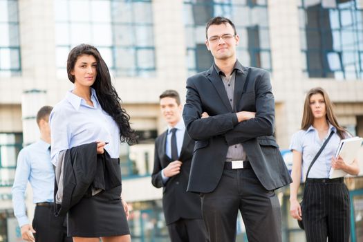 Young businesswoman and businessman with his team in the background standing in front of office building, looking at camera.
