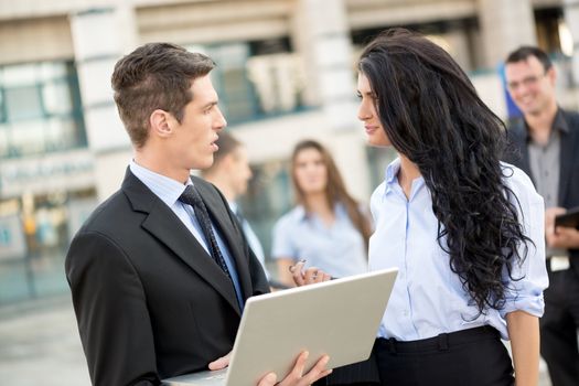 Young businessman talking to his business partner in front of office building holding a laptop while in the background see the rest of the business team.