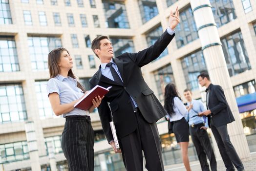 Handsome businessman and his secretary standing in front of office building separated from the rest of the business team. Businessman carrying in one hand a closed laptop while his other hand pointing to the heights, and the secretary keeps the organiser which records the bosses idea.