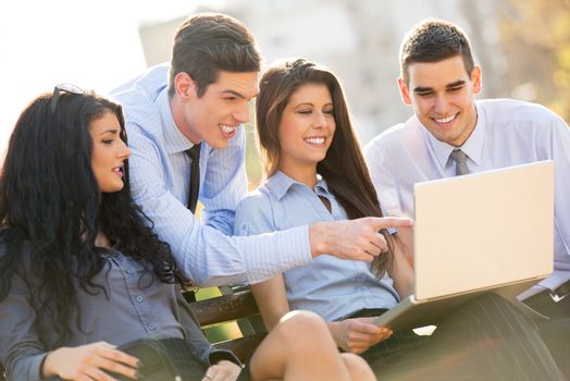 A small group of young business people sitting on a park bench during a break looking at the laptop.