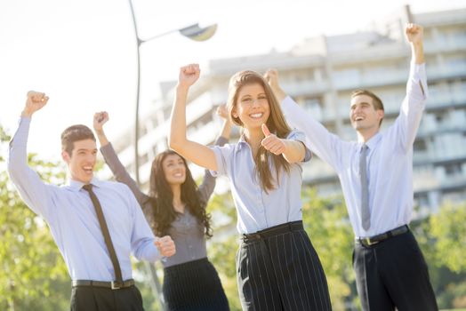 Young businesswoman elegantly dressed with his team celebrating business success at a park near his office building, arms raised and outstretched thumb on looking at the camera.