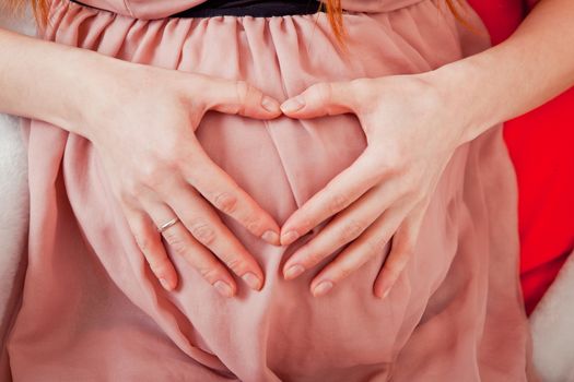 Pregnant Woman holding her hands in a heart shape on her baby bump. Pregnant Belly with fingers Heart symbol. Maternity concept. Baby Shower.