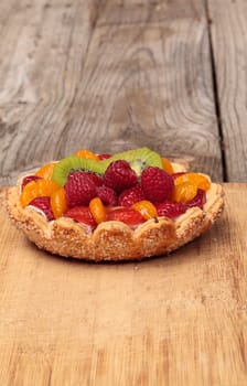 Fruit tart with a yellow custard, ladyfinger cookie shell crust, raspberries, strawberries, kiwi fruit and mandarin oranges on a cutting board on a rustic farm picnic table in summer.