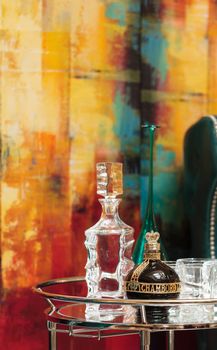 Orange, CA, USA — April 29, 2016: Alcohol decanter, bottle of Chambord liqueur, red wine and glasses on a glass serving table between two chic blue green leather chairs in front of a red, orange and yellow abstract painting.