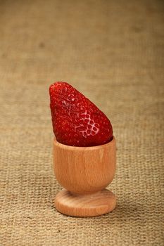 One big red mellow strawberry in wooden eggcup holder on natural burlap jute canvas background