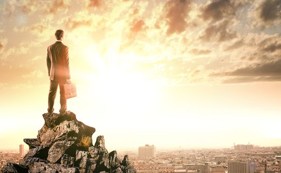 Young businessman standing on edge of rock mountain and looking at city