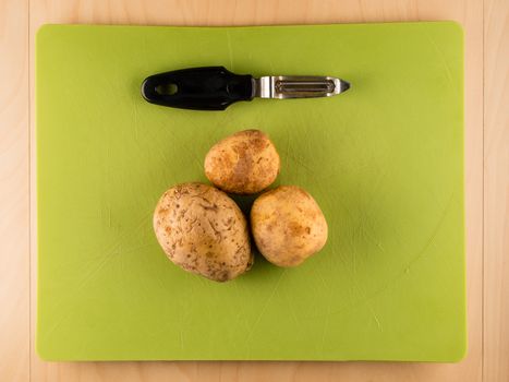 Several three unpeeled potatoes on green plastic board with peeler, simple food preparation illustration, vegetarian dieting, top view still life with center composition