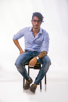 Portrait of brunette young man in light blue shirt and jeans, sitting in studio shot against light background. Full length photo