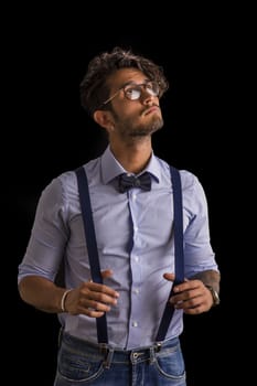 Portrait of brunette young man in glasses, bow-tie, suspenders and shirt looking at camera. Studio shot isolated on black background