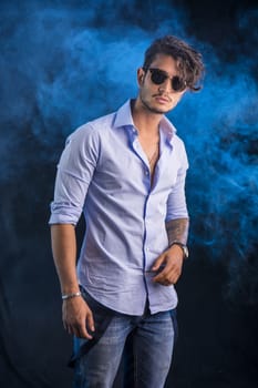 Portrait of brunette young man in light blue shirt and jeans, standing in studio shot against dark smoky background