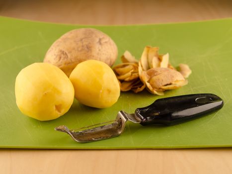 Two peeled and one unpeeled potatoes on used green plastic board with peeler, simple food preparation illustration, vegetarian dieting, still life