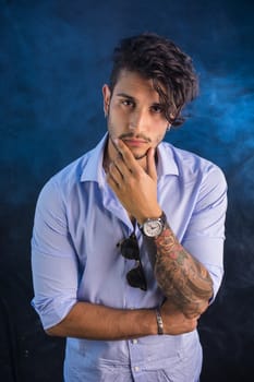 Portrait of brunette young man in light blue shirt and jeans, standing in studio shot against dark smoky background