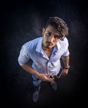 Portrait of brunette young man in light blue shirt and jeans, standing in studio shot from above perspective, against dark background. Full length photo