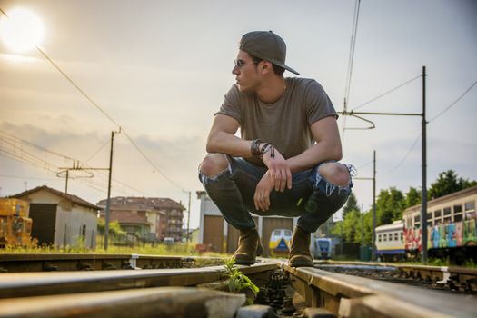Attractive young man sitting on railroad, wearing grey t-shirt and jeans, looking away