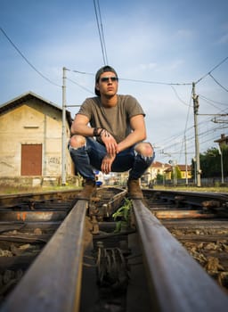 Attractive young man sitting on railroad, wearing grey t-shirt and jeans, looking away