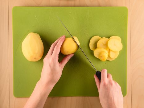 Hands holding knife and slicing potatoe on green plastic board, simple food preparation illustration, vegetarian dieting, top view still life with center composition