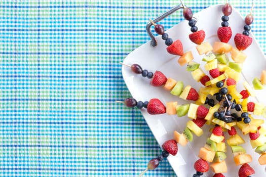 Fresh healthy tropical fruit shish kebabs arranged neatly on a tray placed on a picnic table with a blue checked design and copy space, viewed from above