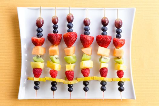 Healthy colorful fresh shish kebab fruit treats made from seasonal summer tropical fruit arranged neatly in a row on a modern white rectangular platter on a yellow textured table viewed from above