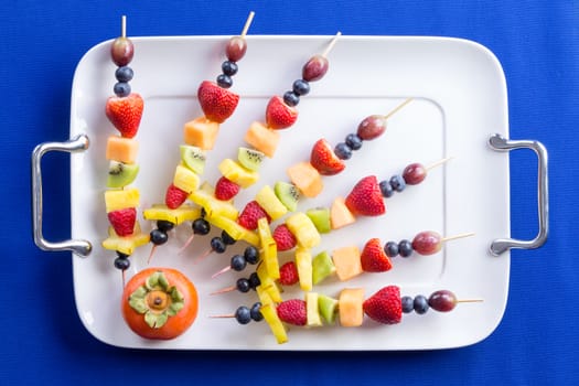 Creative arrangement of colorful fruit kebabs with tropical fruit on skewers radiating from the corner of a tray with a ripe juicy persimmon, overhead view on a blue background