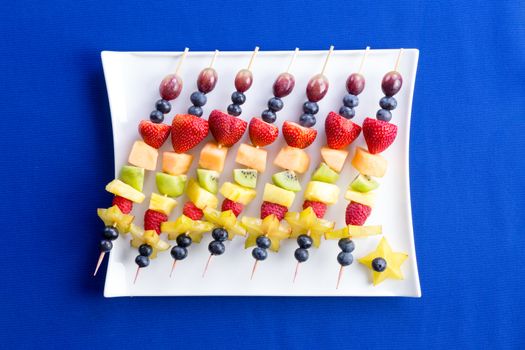 Healthy fruit kebabs with tropical seasonal summer fruit arranged diagonally on a modern rectangular plate over a texture blue background, viewed from above