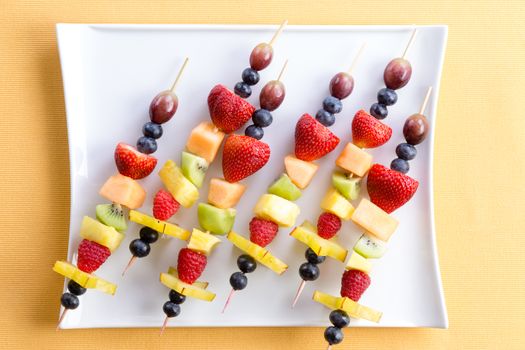 Fresh fruit shish kebabs arranged diagonally on a modern square plate for a healthy vegetarian buffet or dessert, viewed from above on a yellow summer table