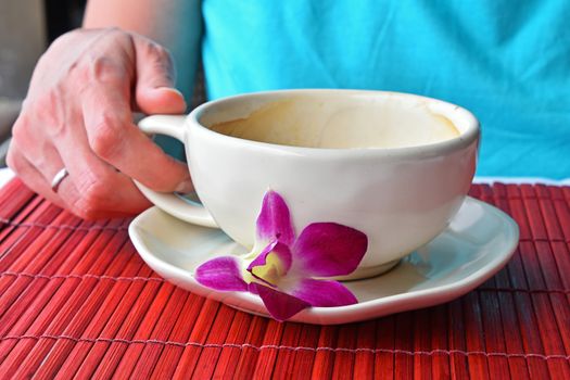 Woman hand holding half empty latte coffee cup with orchid flower at porcelain saucer