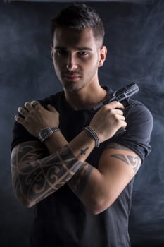 Young handsome man holding a hand gun, wearing black t-shirt, arms crossed on chest, on dark background in studio