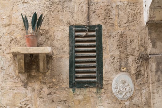 Detail of a wall of an old house in Senglea, island Malta. Pot with iron green leaves, window and a saint picture. Structured facade.