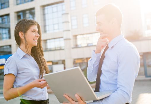 Young businesswoman and her colleague with a laptop stand in front of their company with a smile on their faces talking to each other illuminated by the light of the setting sun.