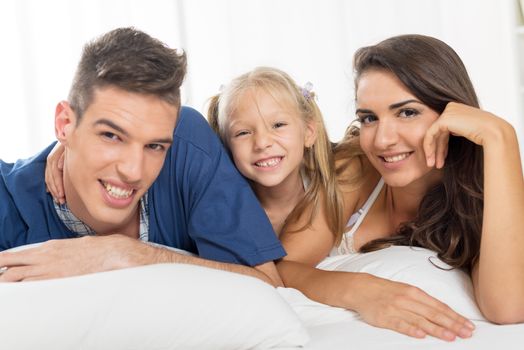 Young parents with their sweet daughter lying in bed looking at the camera with a smile.