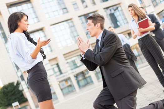 Young handsome, elegantly dressed businessman on the street in front of office building, kneeling with folded hands in front of a young attractive business woman while they watching colleagues.