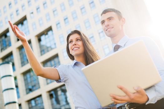 Young business couple with laptop, standing in front their company and look ahead at the height illuminated by the backlight of the sun's rays.