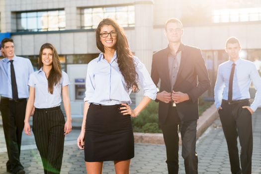 Beautiful young businesswoman with glasses standing proudly with his young colleagues in front of the office building lit by the rays of the setting sun.