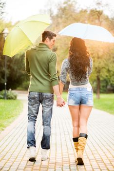 Loving couple, photographed from behind, walking through the park on a rainy day holding hands and carrying umbrellas.