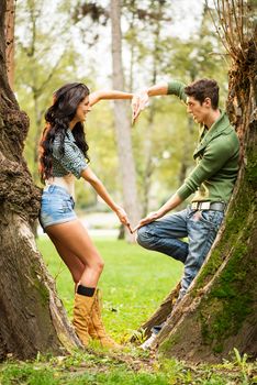 Young and good-looking girl and guy at park leaning against a tree trunk, facing each other with their hands straight form the heart.