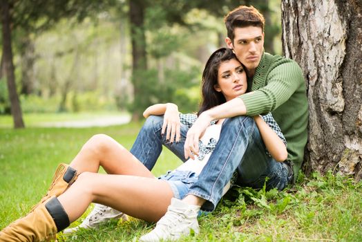 Young handsome guy and attractive leggy girl sitting on the grass in the park. The young man was leaning against a tree, and the girl on the boy between his legs in a half-sitting position.