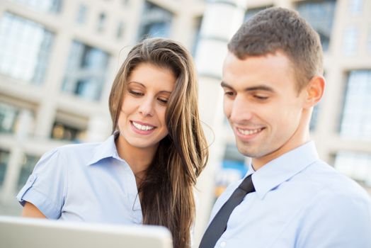 Portrait of a young business couple while standing in front of office building, looking at laptop.