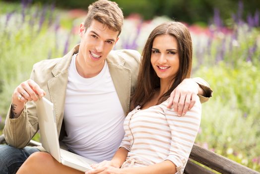 Young heterosexual couple sitting on a park bench enjoying a beautiful sunny day and smiling looking at the camera while a girl holds a laptop in her lap.