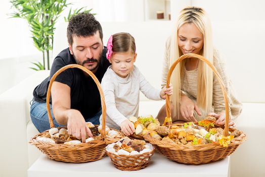 Happy family together, parents with daughter sitting on the couch in the living room in front of woven baskets filled with pastry, choosing the next snack.