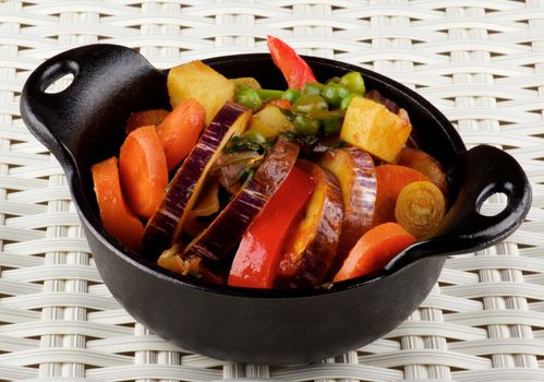 Delicious Homemade Colorful Vegetables Ragout with Striped Eggplant, Carrots, Potatoes, Leek, Red Bell Pepper and Green Pea in Black Iron Stewpot closeup on Wicker background