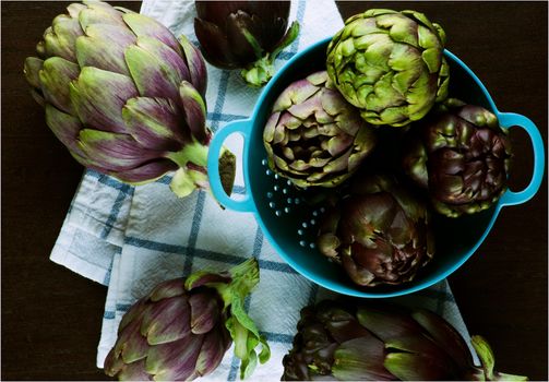 Heap of Big and Small Raw Artichokes in Blue Colander on Checkered Napkin closeup on Dark Wooden background. Top View