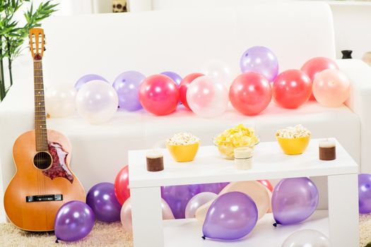 Room with a couch where is leaned acoustic guitar, couch and floor are covered with balloons, and on the table in front of the couch, there are decorative candles and snacks.