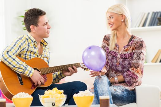 Young heterosexual couple sitting on a couch in the room, a young man playing an acoustic guitar seduces a pretty girl, which holds balloon in hand.