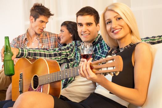 Small group of happy young people hang out at the house party with an acoustic guitar.