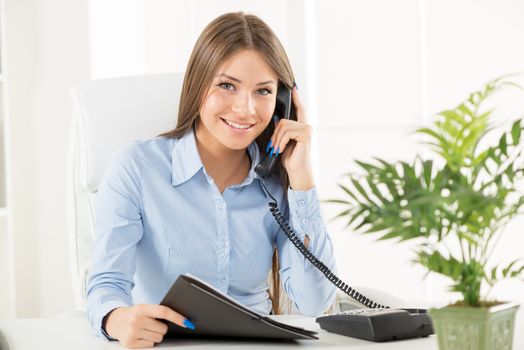 Young business woman phoning in office, sitting at an office desk with one hand holding the phone, and in another folder.